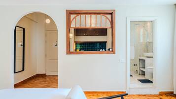 A modern hotel room with a wooden floor, a large bed, a built-in window with a wooden frame, and a separate bathroom with a glass shower enclosure.