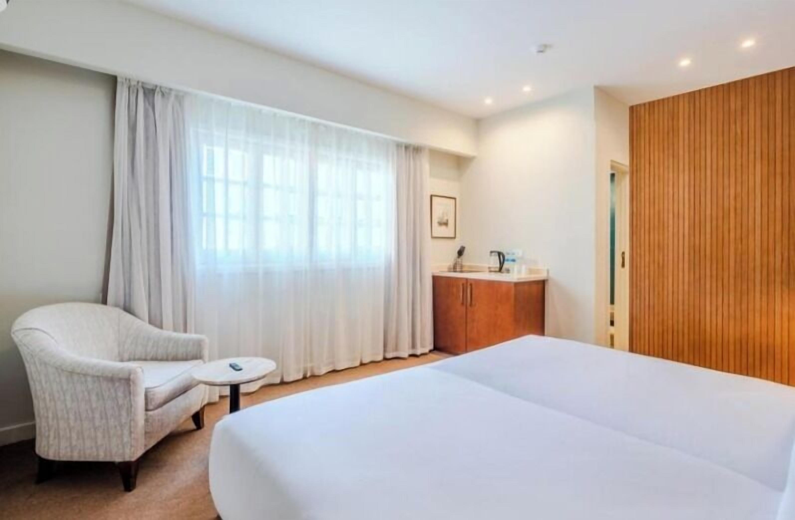 A hotel room with a large bed, a chair, a small table, a wooden wardrobe, and a window with curtains.