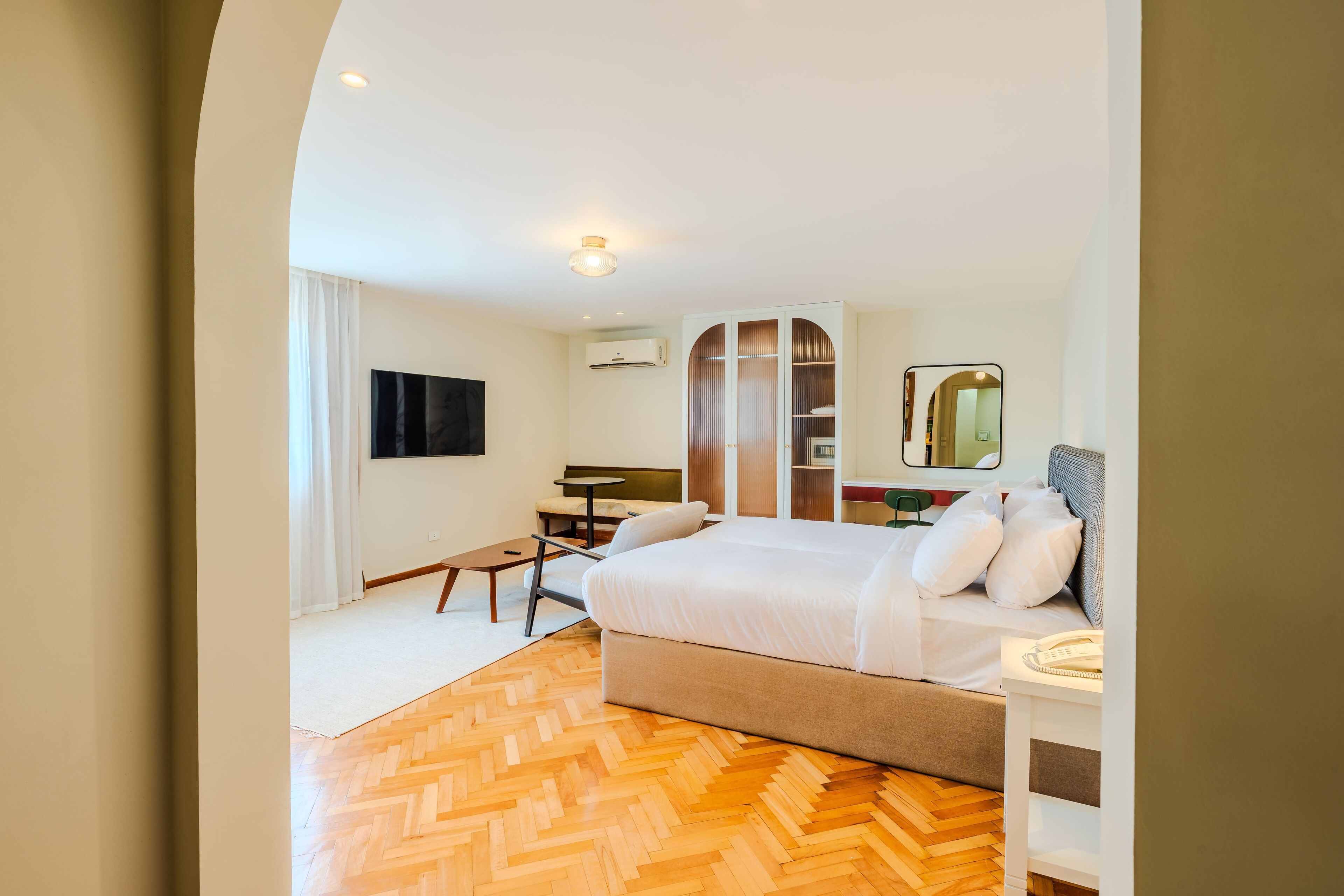 A hotel room with a bed, bedside table, and a small desk with a chair. There is a wardrobe with a mirror and a window with curtains.
