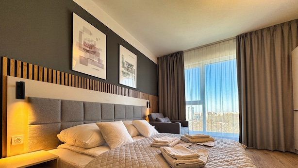 A modern hotel room with a large bed, a bedside table, a chair, and a view of the city through large windows. - Warszawa, Wolska 2-3 osobowy apartament z klimatyzacją (Warszawa)