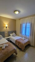 Habitación de hotel con dos camas, una mesita de noche, una lámpara y una ventana con cortinas translúcidas.