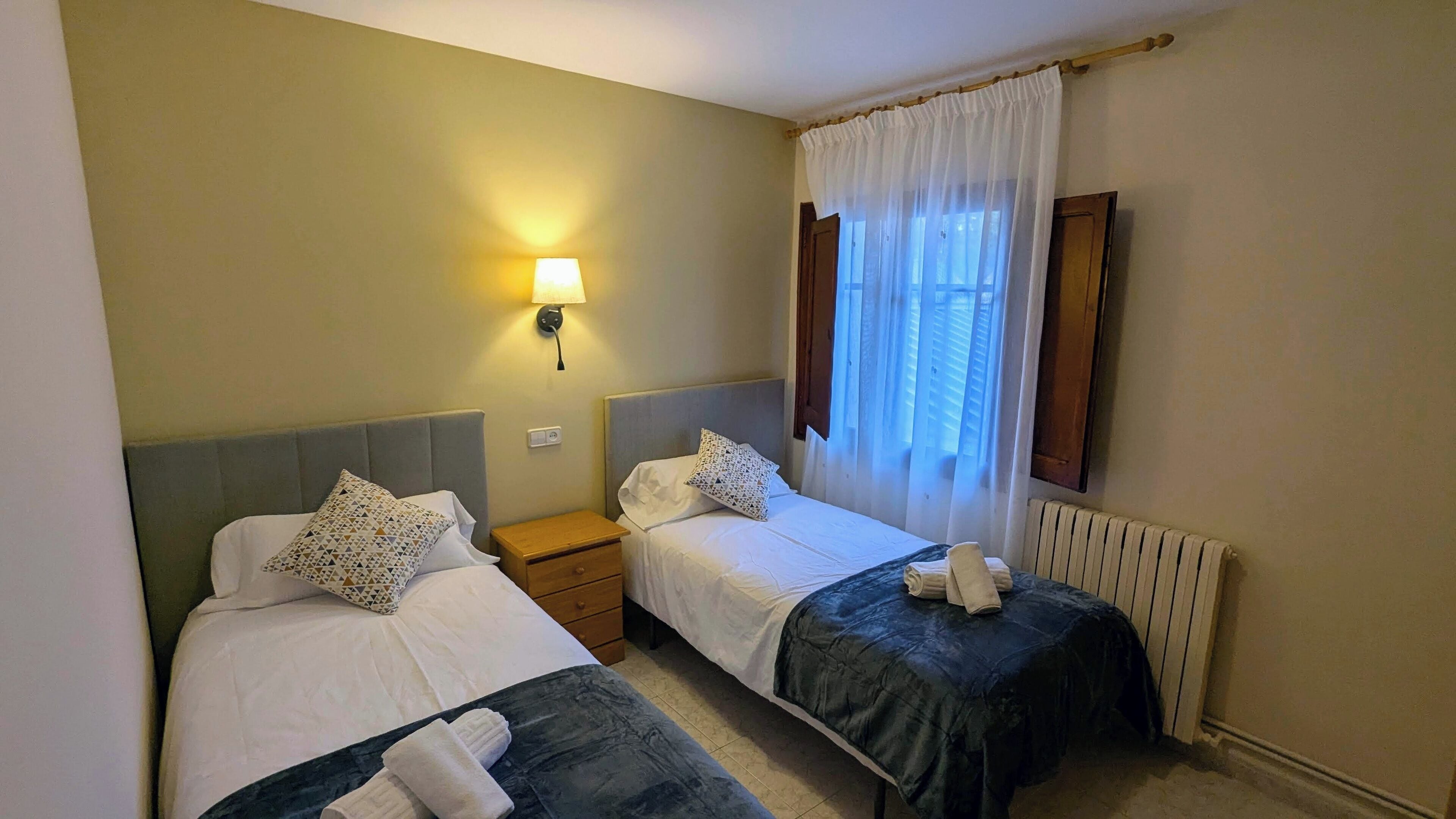 Habitación de hotel con dos camas individuales, una mesita de noche, una lámpara, una ventana con cortinas y un radiador.