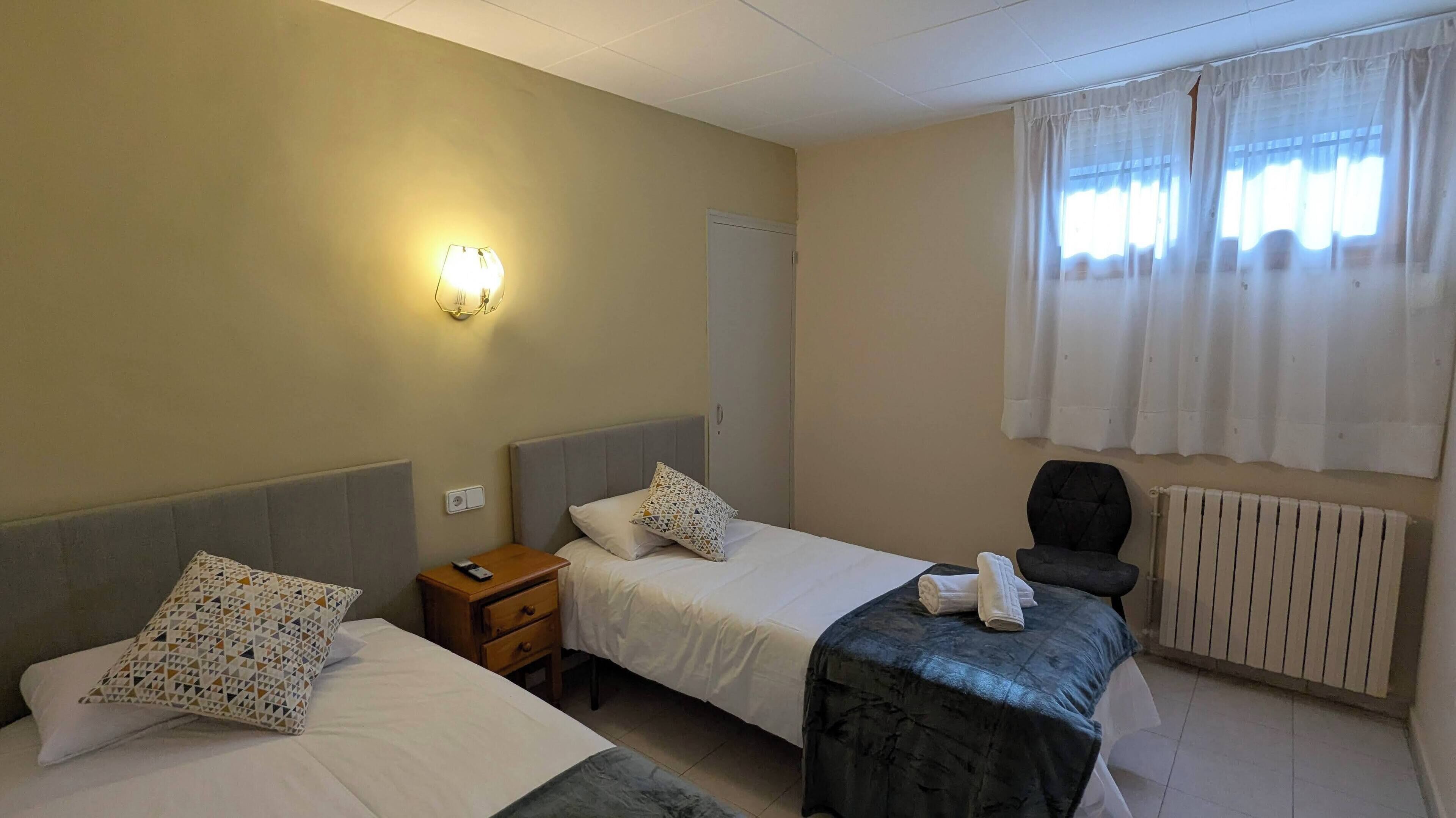 A hotel room with two beds, a bedside table with a lamp, a chair, and a radiator.
