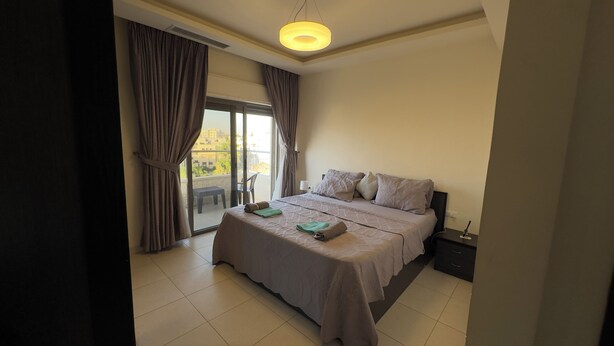 A hotel room with a bed, a nightstand, a lamp, and a view of the outside. - Modern fully furnished 4-Room apartment (Amman)