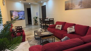 A living room with a red sofa, a coffee table, and a TV displaying a cityscape. There is a dining area with a table and chairs, and a door leading to another room. - Modern fully furnished 4-Room apartment (Amman)