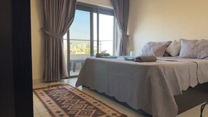 A hotel room with a bed, a view of the city through large windows, and a patterned rug. - Modern fully furnished 4-Room apartment (Amman)