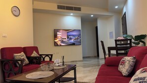 A living room with a TV displaying a cityscape, red sofas, a wooden coffee table, and a dining area with a chair and table. - Modern fully furnished 4-Room apartment (Amman)