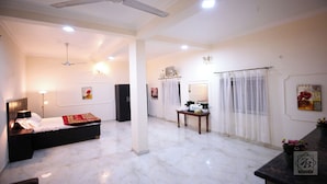 Une chambre d’hôtel comprenant un lit, un bureau, une chaise, un ventilateur de plafond, une fenêtre avec des rideaux et des œuvres d’art accrochées aux murs.