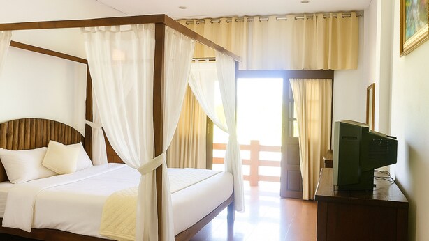 Uma cama com dossel, roupa de cama branca, cabeceira de madeira e cortinas leves.