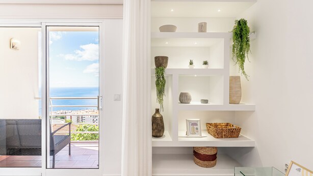En moderne stue med balkon, hvide reoler med pynteting, og havudsigt.