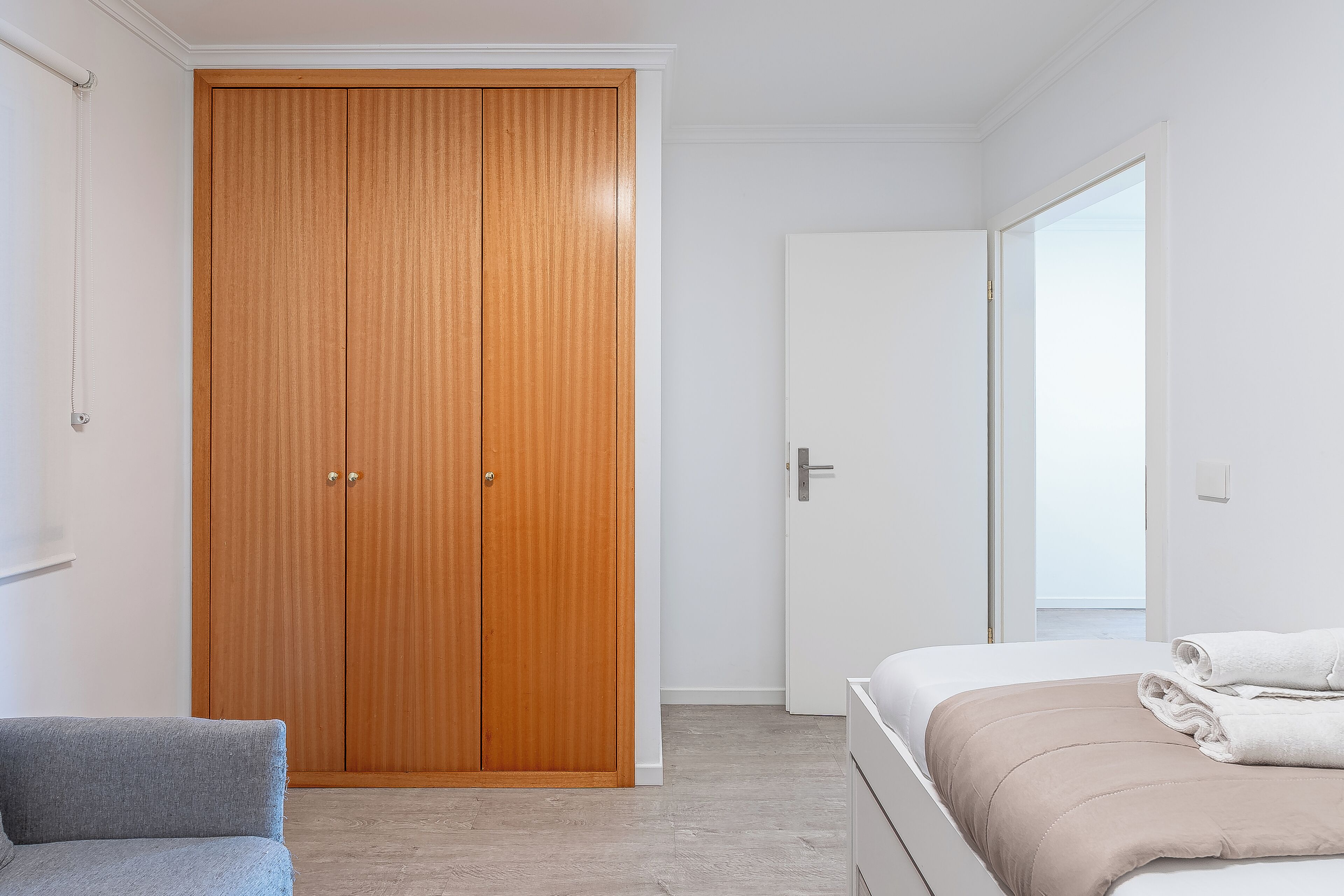 Un dormitorio con un armario de madera, una cama con una manta color beige y un sillón gris.
