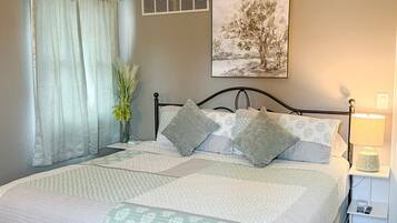 Un lit bien fait, recouvert d’une couvre-lit à motifs, une table de chevet avec une lampe et une bouche d’aération fixée au mur.