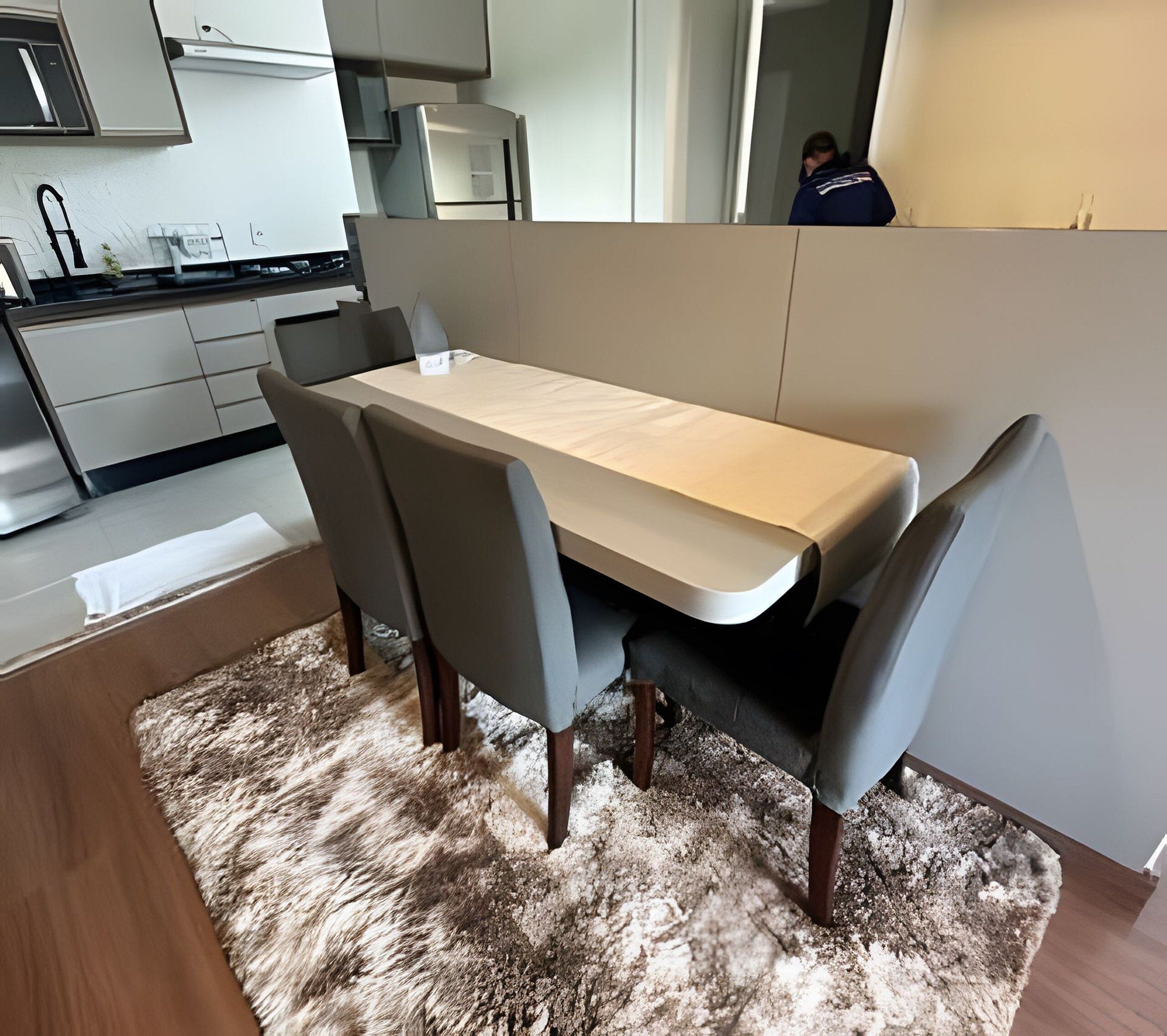 A modern dining area with a wooden table and grey chairs, a fluffy rug, and a kitchen in the background.