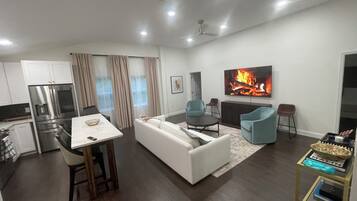 A modern living room with a white sofa, two blue armchairs, a dining table with chairs, a kitchen area with a refrigerator, and a flat-screen TV displaying a fireplace.