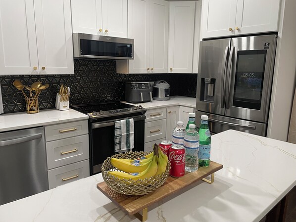 A modern kitchen with white cabinets, a stainless steel refrigerator, a microwave, and a stove. There are bananas, a can of Coca-Cola, and bottles of water on the counter.