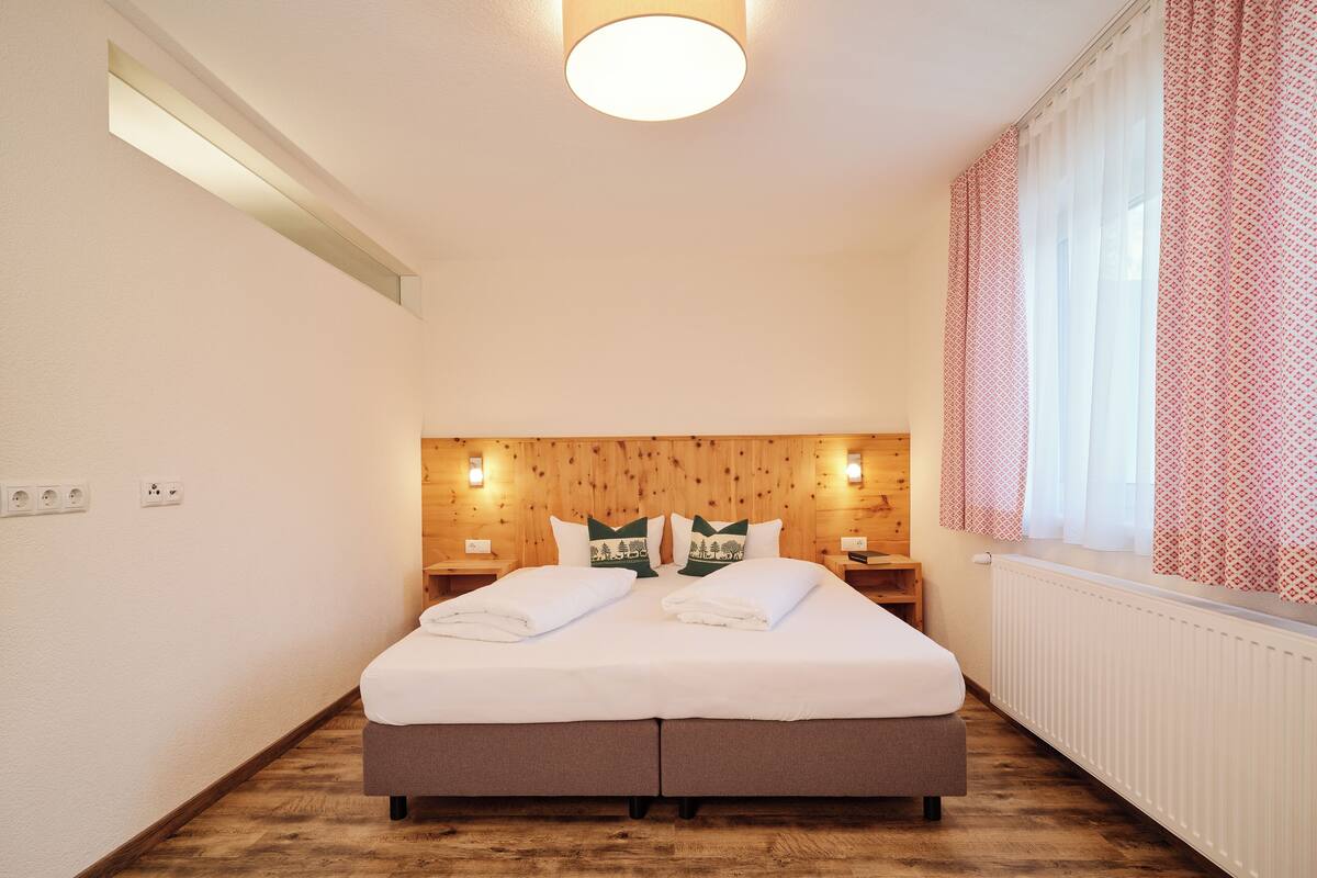Ein Doppelbett mit hölzernem Kopfteil, zwei Nachttische und ein Fenster mit gemusterten Vorhängen.