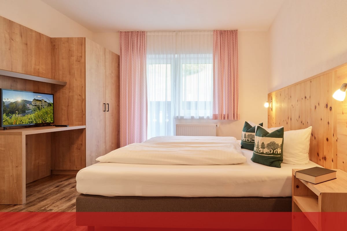 Ein Hotelzimmer mit einem hölzernen Kopfteil, einem Bett mit weißer Bettwäsche, einem Fernseher auf einer Holzablage und einem Fenster mit durchsichtigen Vorhängen.