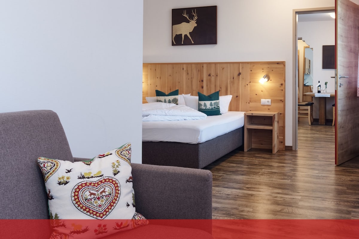 Ein gemütliches Zimmer mit einem Bett, einem hölzernen Kopfteil, einem Nachttisch und einem Sessel mit einem gemusterten Kissen.