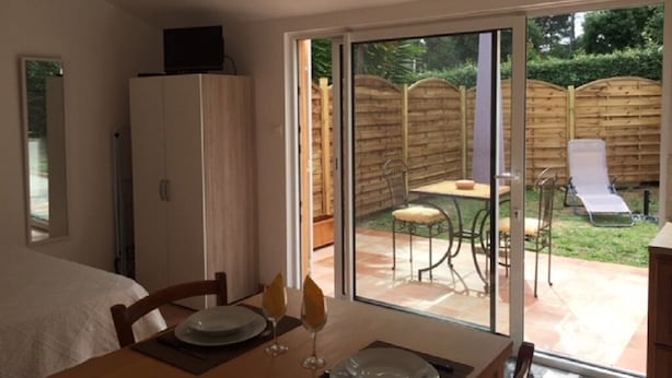 A dining area with a table set for two, a sliding glass door leading to a patio with a table and chairs, and a white cabinet with a television on top.