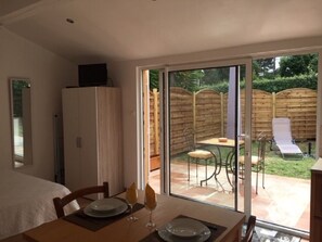 A dining area with a table set for two, a sliding glass door leading to a patio with a table and chairs, and a white cabinet with a television on top. - Separate studio with a wooded garden, near shops and beaches (Six-Fours-les-Plages)