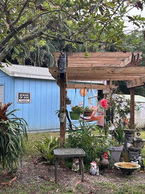 A wooden pergola with a 'Beware of Dog' sign, surrounded by potted plants, a hammock, and various garden decorations. - The Funky Chicken studio is unique. Pet friendly. Key West atmosphere. (Naples)