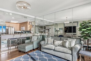A modern living room with a grey sofa, a coffee table, and a large mirror reflecting the kitchen area. - Stylish 17th-Floor Condo with Stunning Gulf Views (Panama City Beach)