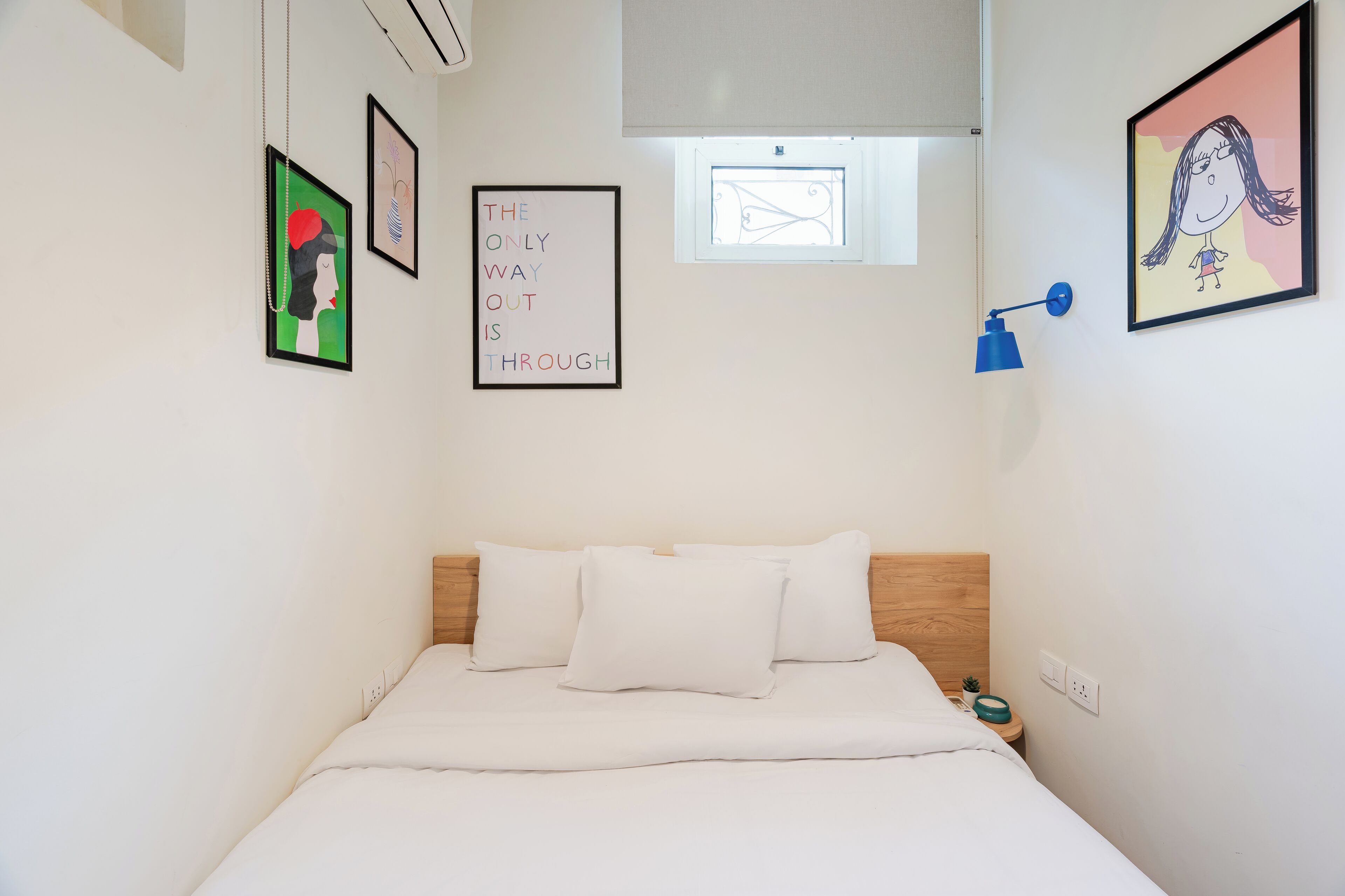 A bedroom with a bed, white bedding, a wooden headboard, a wall-mounted lamp, and framed artwork.