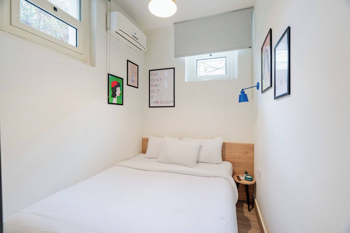 Una habitación pequeña y limpia con cama, una mesita y aire acondicionado de pared.