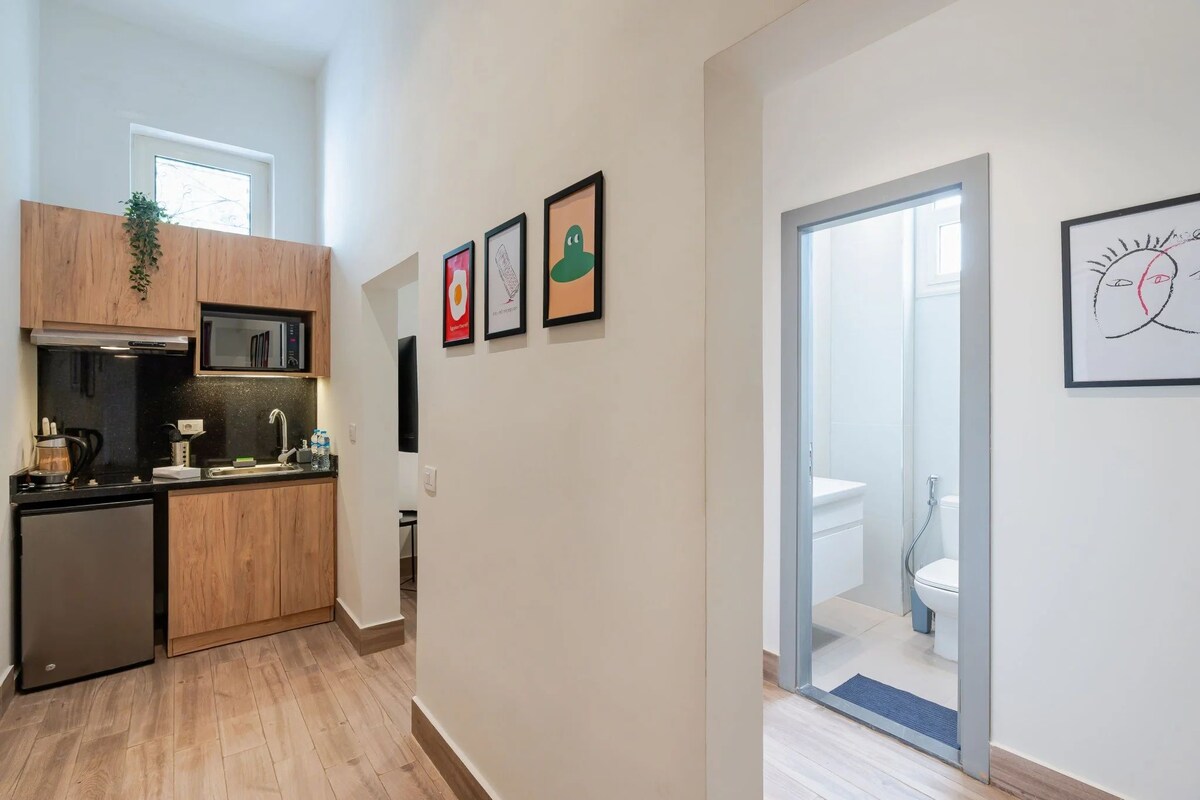 Een moderne keuken met houten kasten, een ingebouwde magnetron en een koelkast. Er hangen kunstwerken aan de muur en er is uitzicht op de badkamer via een glazen deur.