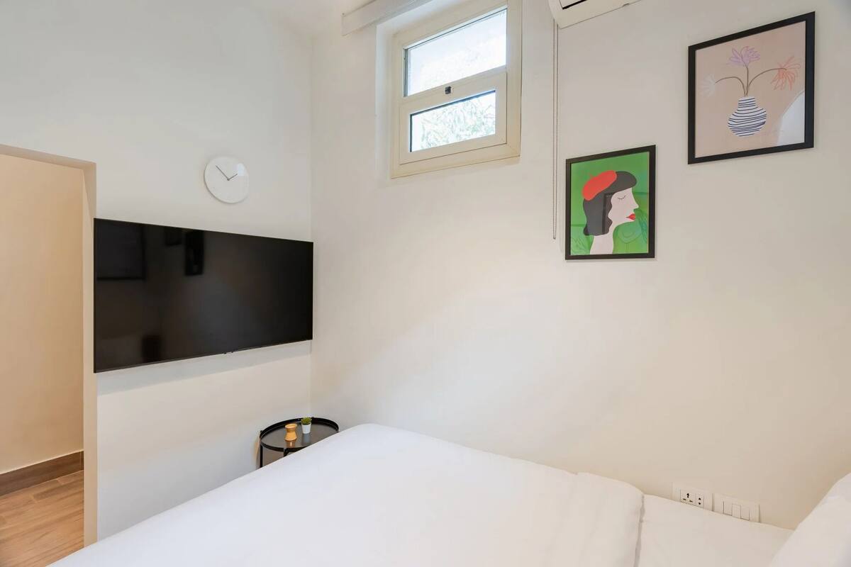 Un dormitorio con una cama, un televisor montado en la pared, un reloj, una ventana y dos cuadros enmarcados.