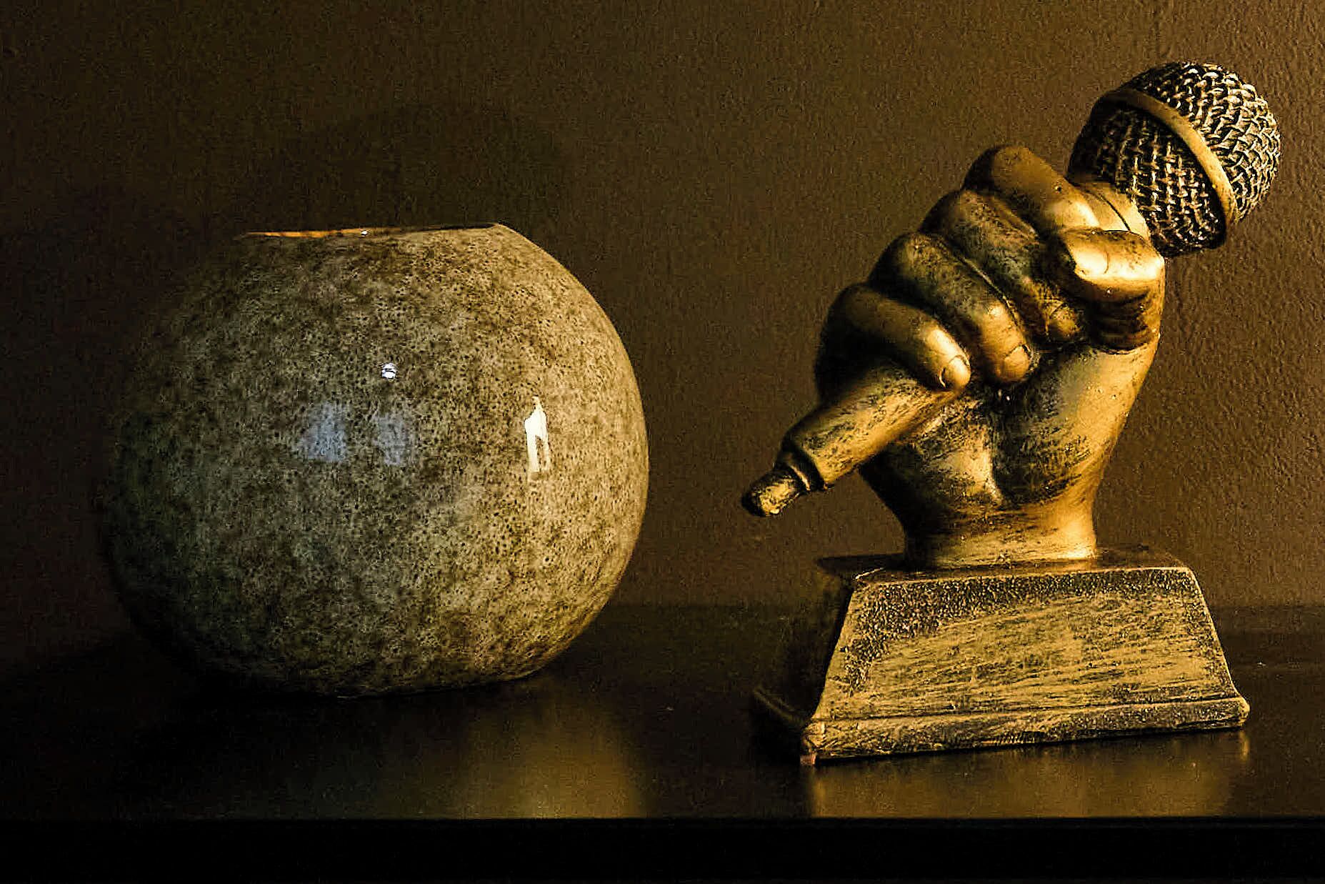 Une sculpture représentant une figure tenant un microphone, placée sur un piédestal à côté d’un vase en pierre.