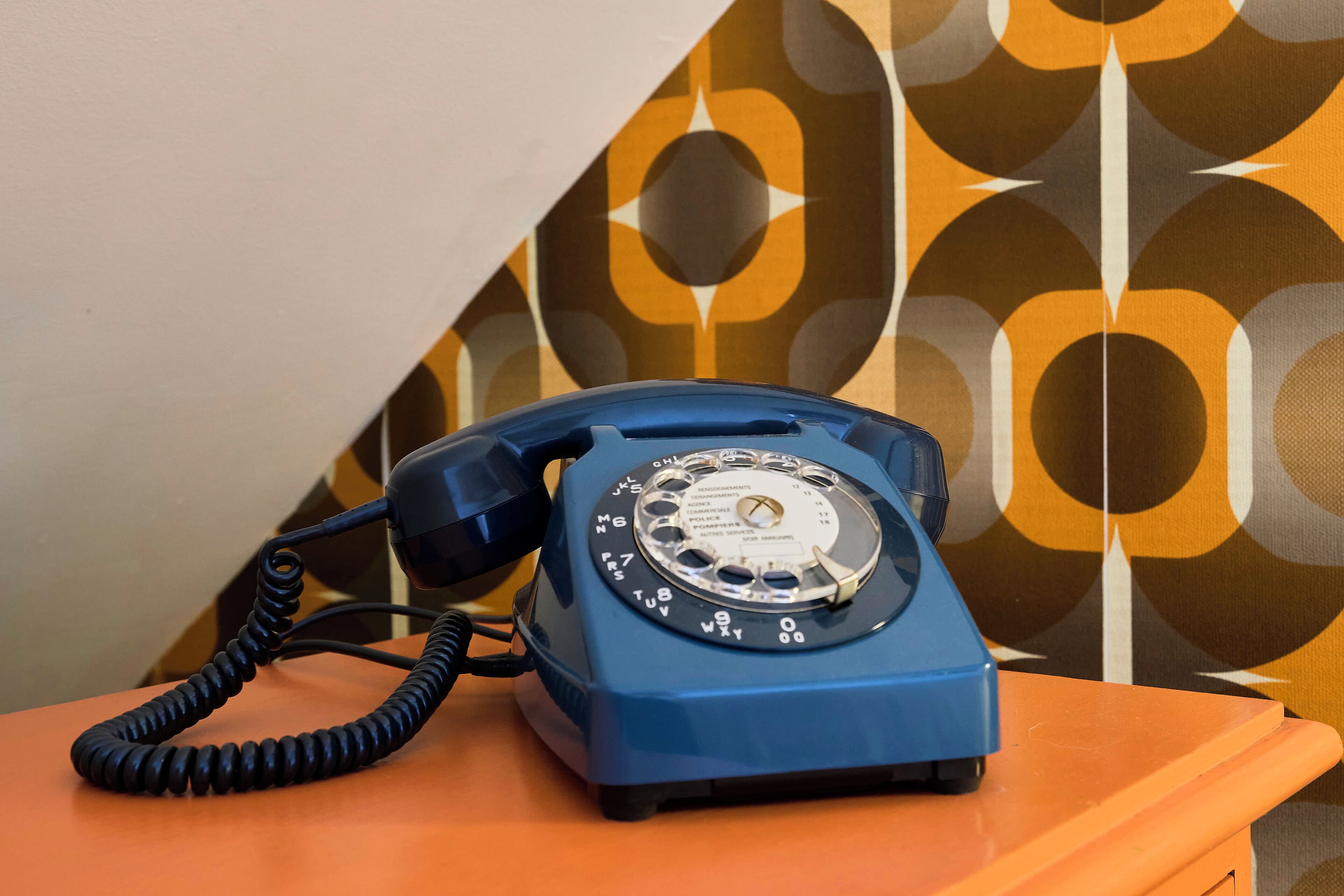 Un téléphone à cadran bleu est posé sur une table orange, devant un mur à motifs.