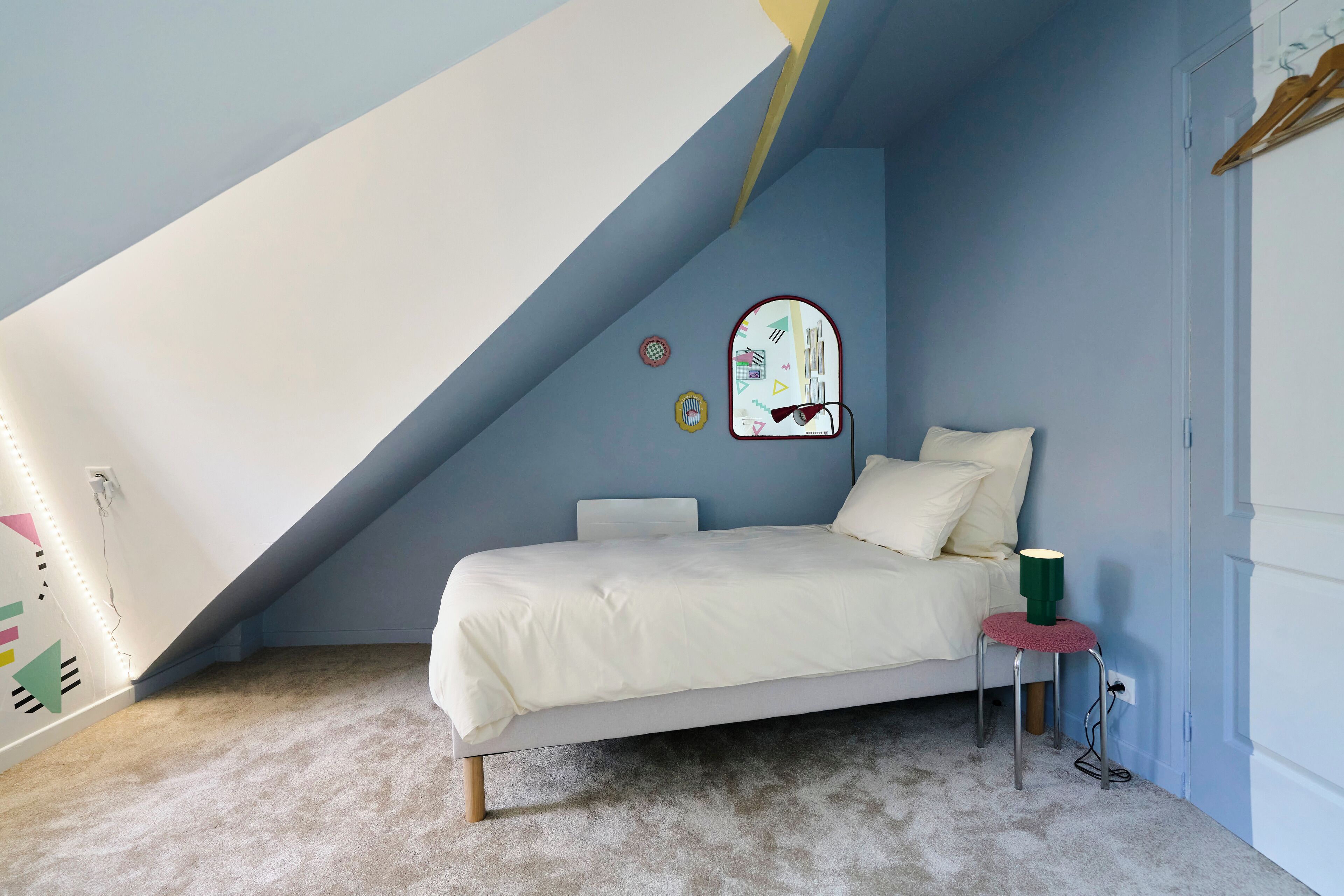 Een slaapkamer met een schuine kap, een bed met witte beddengoed, een klein krukje met een groene lamp, en een spiegel aan de muur.