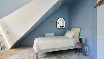 Een slaapkamer met een schuine kap, een bed met witte beddengoed, een klein krukje met een groene lamp, en een spiegel aan de muur.