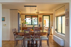 A dining area with a wooden table and chairs, a kitchen in the background, and a window with blinds.