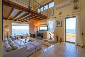 A spacious living area with a large window overlooking the sea, a sofa, a TV, and a fireplace.