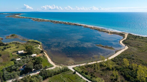 Luftaufnahme einer Küstenlandschaft mit blauem Meer, Sandstrand und einigen Gebäuden, die von Grünanlagen umgeben sind.