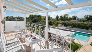 Ein Balkon mit weißen Möbeln, einem Tisch mit Flasche und Gläsern sowie Blick auf einen Swimmingpool und Grünflächen.