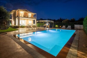 Une piscine agrémentée d’une pergola et de chaises longues, une maison à deux étages dotée d’un éclairage extérieur et un ciel nocturne dégagé.