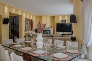 Une salle à manger avec une table en verre dressée pour six personnes, entourée de chaises. On aperçoit une cheminée et un téléviseur à écran plat au fond.