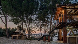 Ein rustikaler Außenbereich mit einer Steinterrasse, einem Backofen aus Ziegeln und einer Holzterrasse mit einer Treppe zu einem überdachten Bereich.