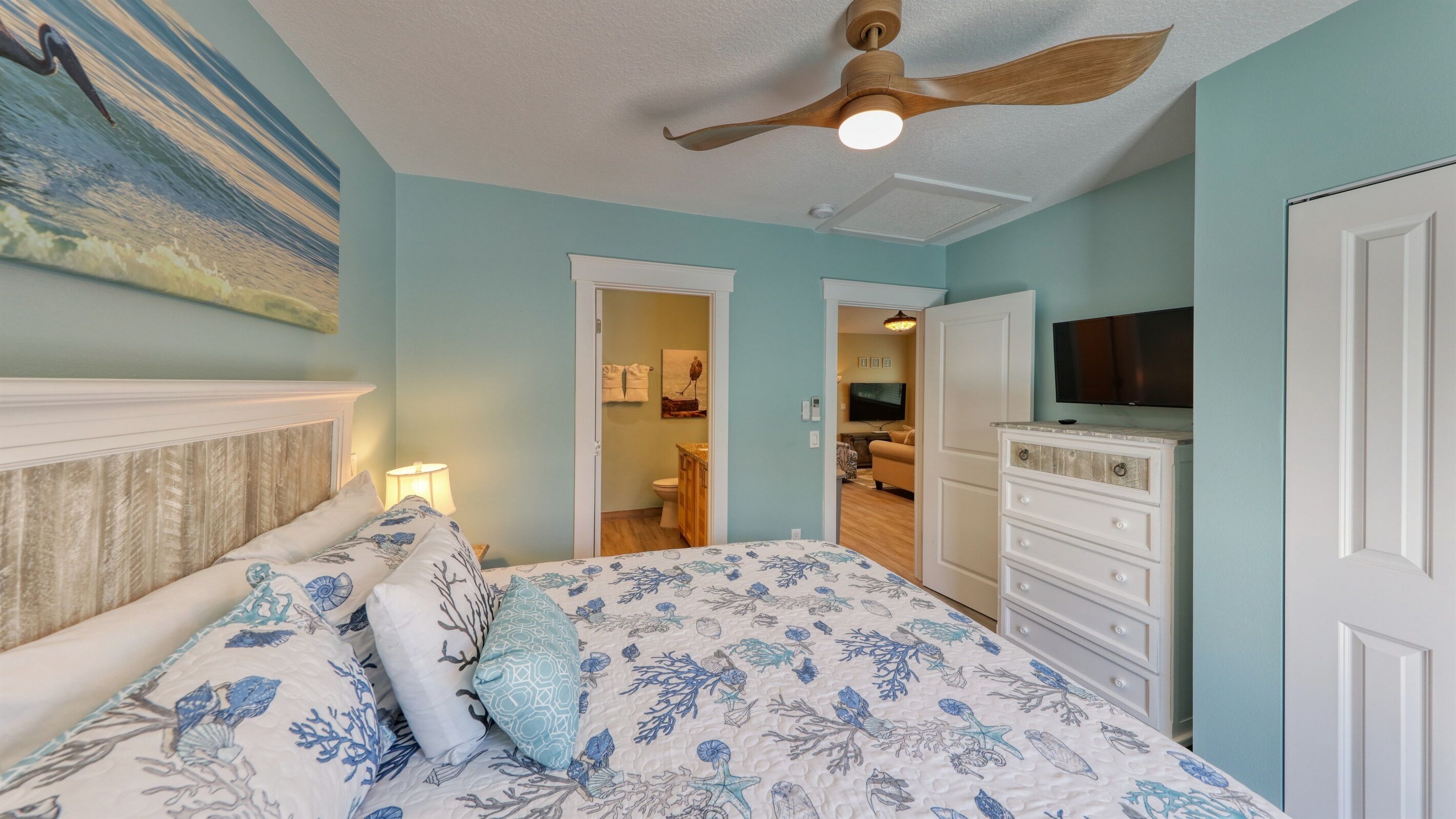 A bedroom with a bed, a ceiling fan, a TV, and a bathroom visible through an open door.