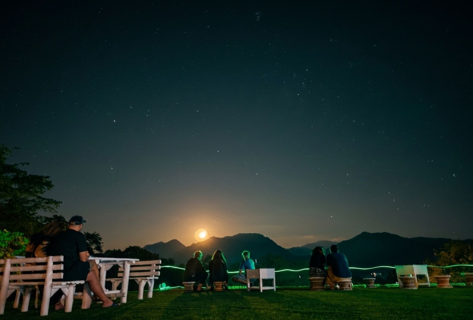 Ein Abendambiente im Freien, eine Gruppe von Personen sitzt auf Bänken unter einem sternenübersäten Himmel, im Hintergrund ein Gebirge.