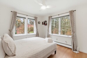 Quarto com cama, duas janelas com cortinas, um ventilador de teto e um relógio na parede.