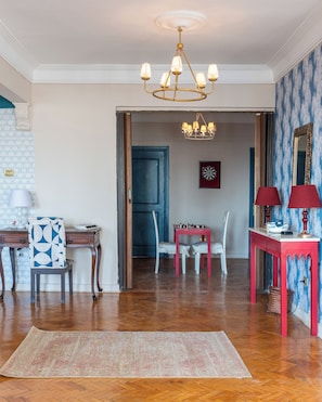 A spacious living room with a wooden floor, a chandelier, a dining area with a red table, and a blue patterned wall. - Zamalek suuny khan (Cairo Governorate)