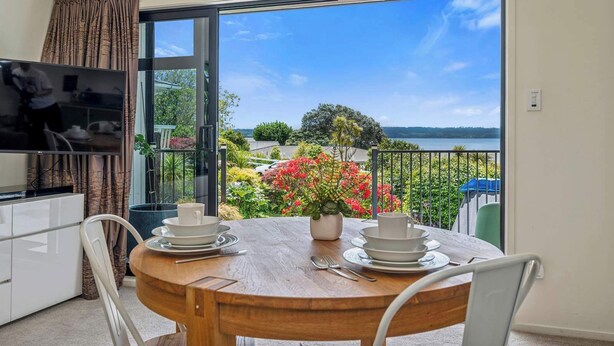 Ein Balkon mit Esstisch für Zwei, einem Fernseher und Blick auf einen See und Grünflächen.