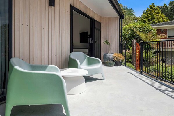 A modern outdoor seating area with two green chairs and a white table, a wooden wall, and a black metal fence. - Inn Manahi - Lake Views, Cosy Retreat for 2 (Rotorua)