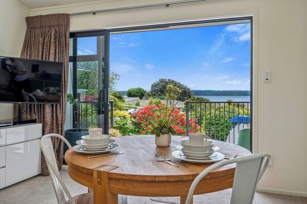 Un balcón con una mesa de comedor para dos personas, un televisor y vistas a un lago y follaje.