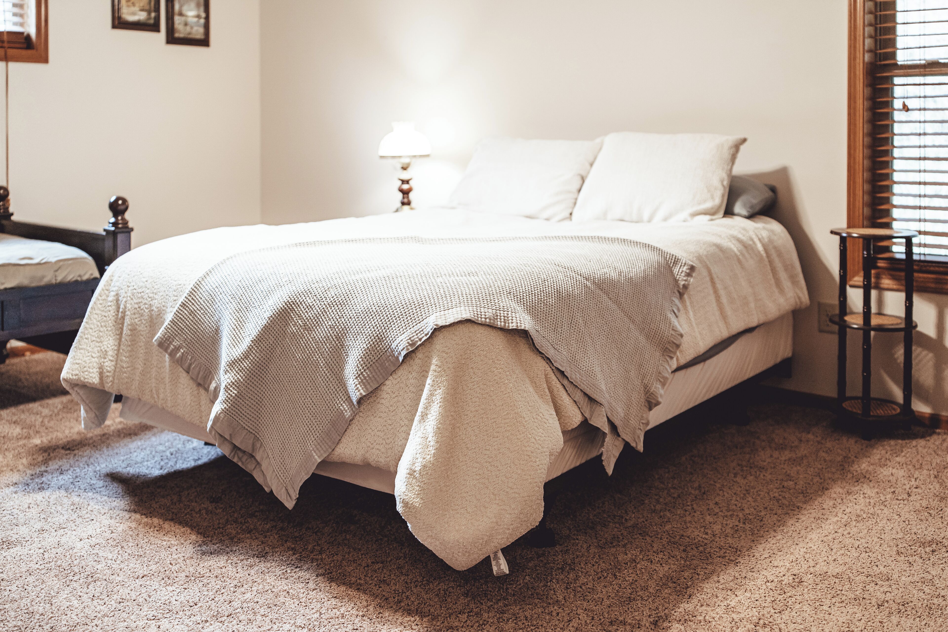 A neatly made bed with a white comforter and pillows, a bedside table with a lamp, and a small wooden stool.