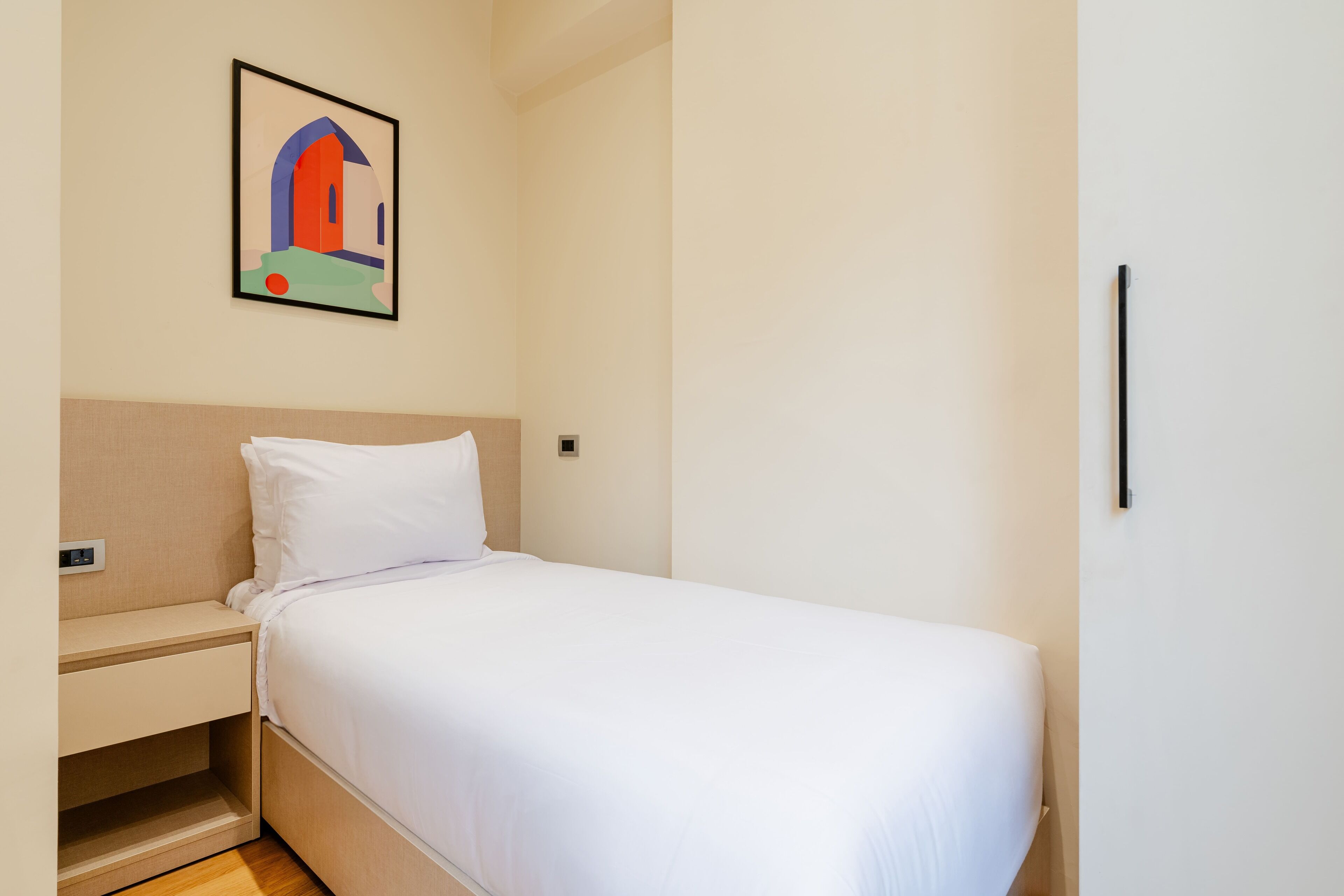 A single bed with white bedding in a room with a beige wall and a framed picture above it.